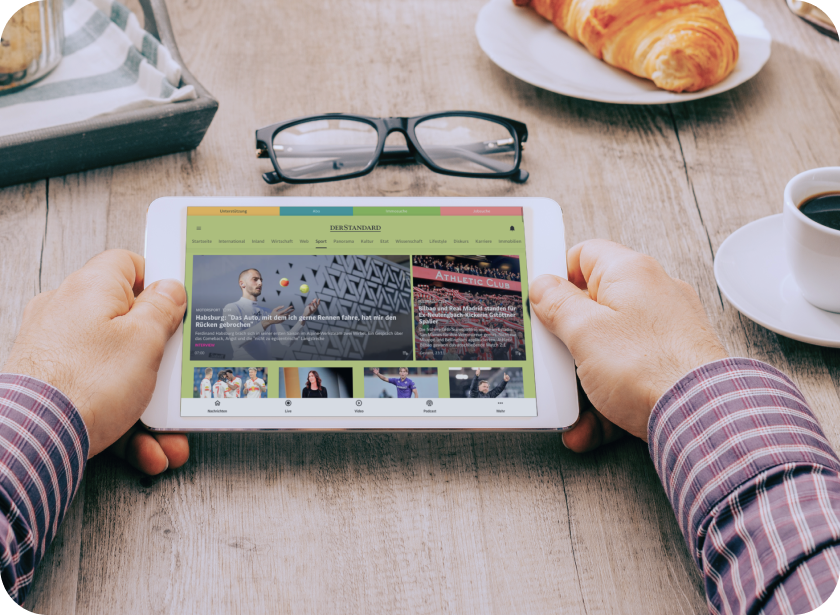 MÃ¤nnerhÃ¤nde, die am FrÃ¼hstÃ¼ckstisch ein Tablet halten, auf dem die DER STANDARD App zu sehen ist.